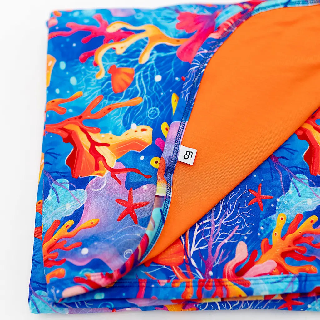 A folded blanket with a vibrant coral, starfish, and sea plant pattern in shades of blue, orange, and purple, with a solid orange underside visible at one end.