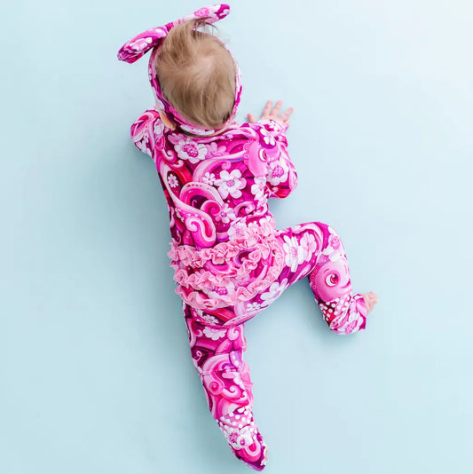 A baby wearing a pink ruffle pajama with a pattern of swirling tentacles and flowers, lying on a light blue background.