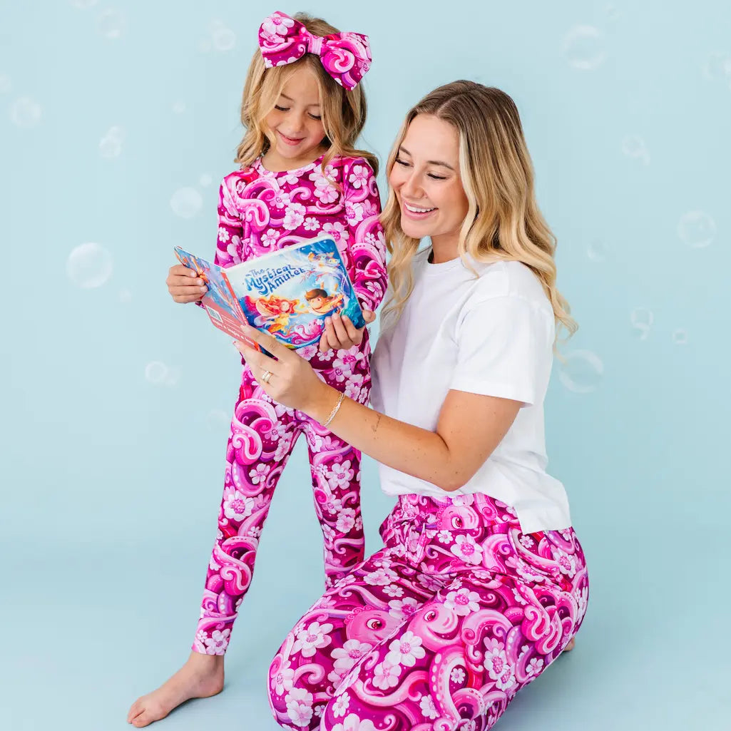 Woman and child in pink and white patterned pajamas holding a book against a light blue background.