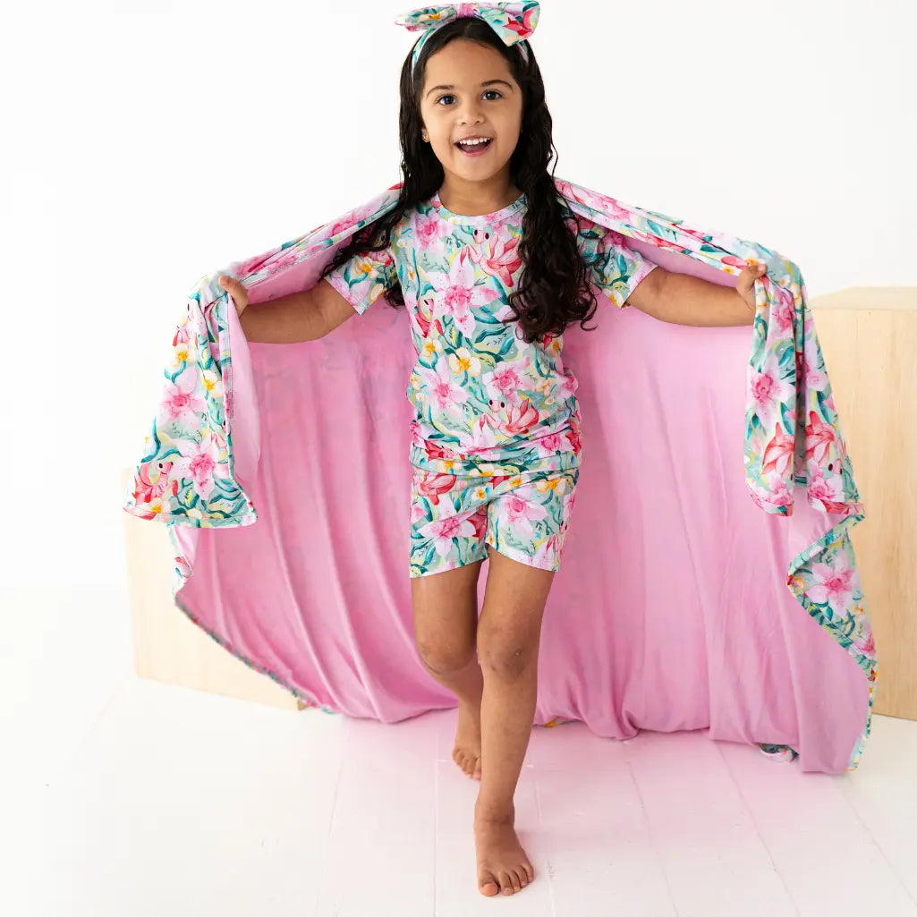 Young girl in floral pajama set with matching cape and headband, standing on a pink blanket.
