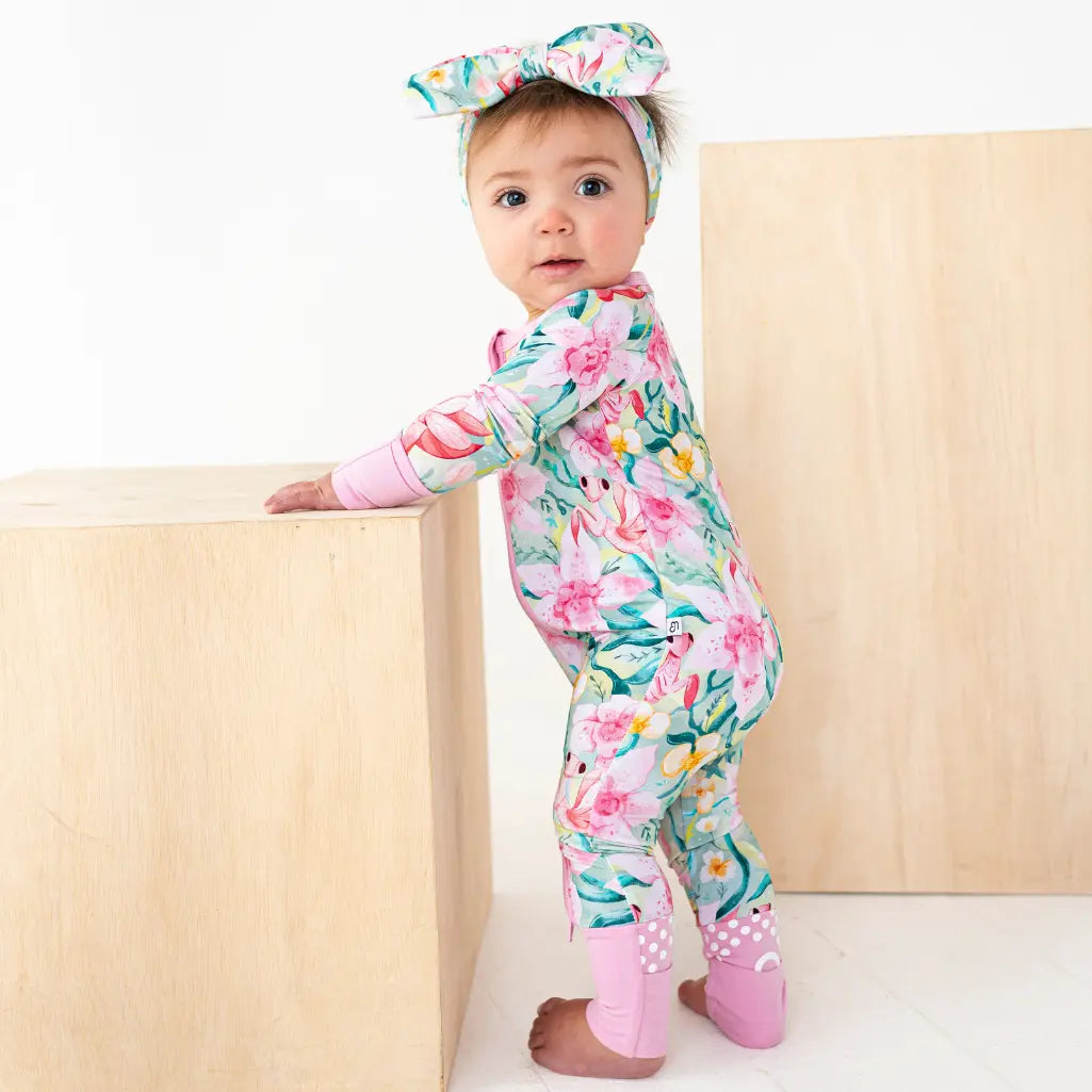 Toddler wearing a pink and teal floral print outfit with matching headband, standing next to a wooden block
