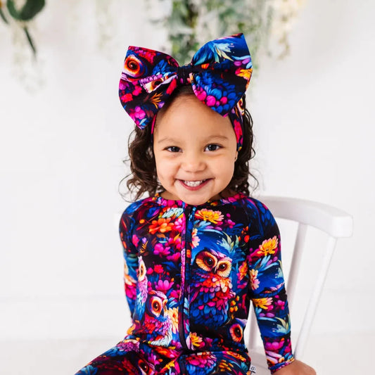 A baby wearing a colorful onesie with owl patterns, including a large bow headband.
