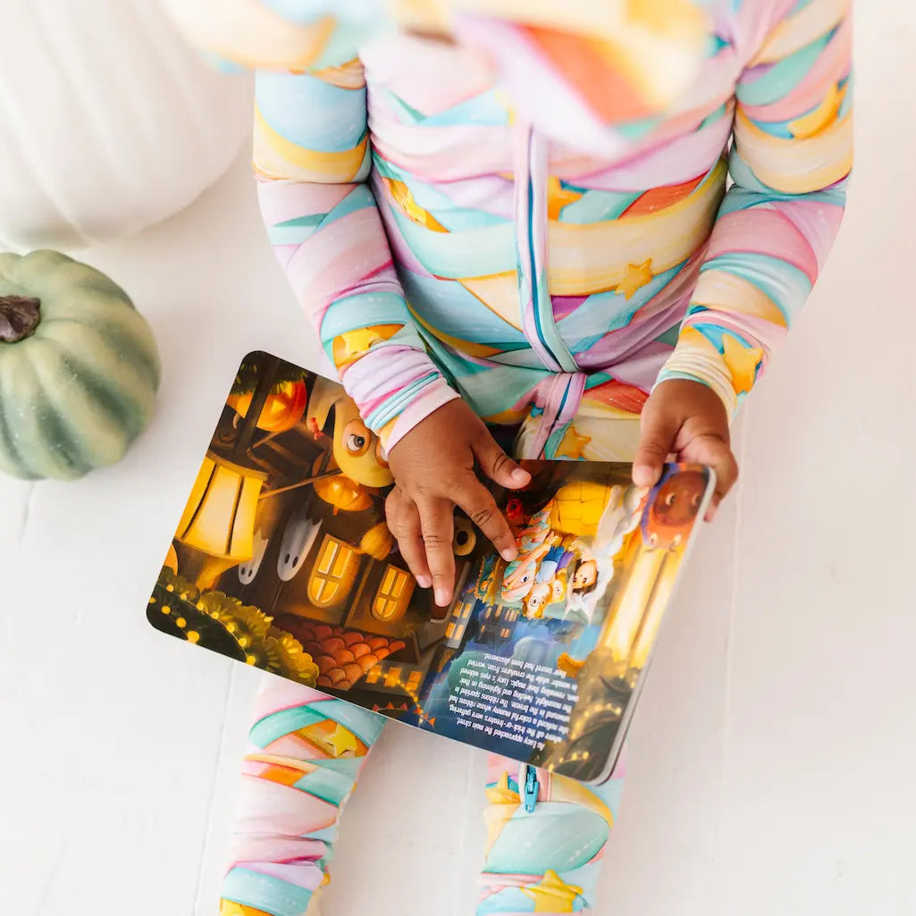 Child wearing colorful pajamas reading a Halloween-themed book with pumpkins on a white surface.