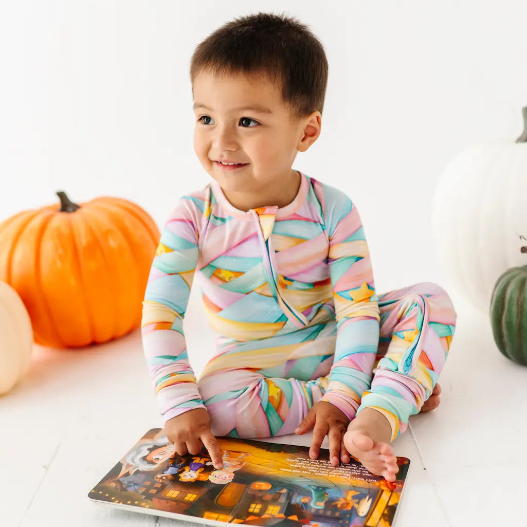 Child wearing colorful pajamas sitting on a white surface with pumpkins and a Halloween-themed book.