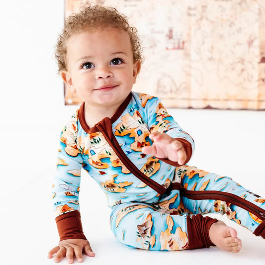Smiling curly-haired toddler sitting on the floor and reaching out, wearing a light blue onesie with an orange map and island print and brown trim, with a treasure-map decoration on the wall behind