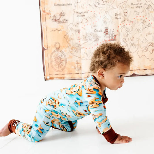 Curly-haired toddler crawling on a white floor wearing a light blue onesie with an orange map and island print and brown trim, with a treasure map decoration hanging on the wall behind