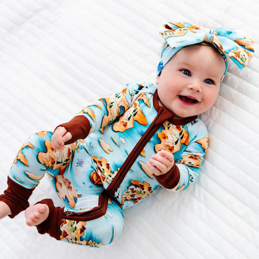 Smiling baby lying on a white blanket wearing a light blue onesie with an orange map and island print and brown trim, plus a matching bow headband