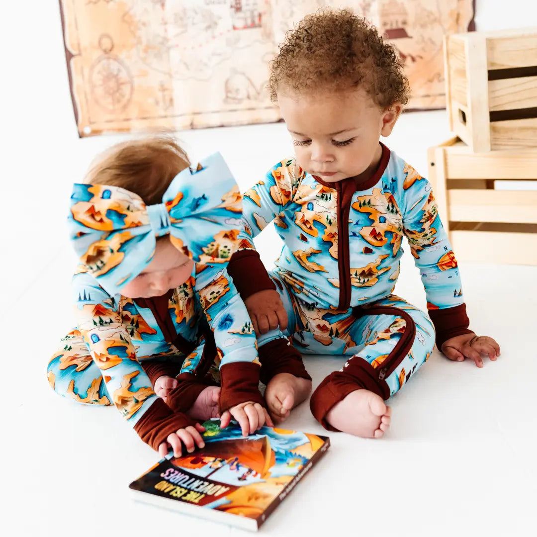 Two children in matching pajamas with a dinosaur pattern, sitting on a bed and looking at a book.
