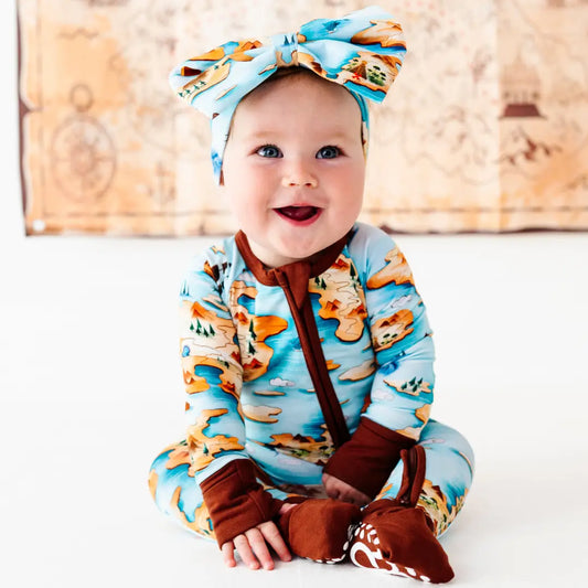 Baby wearing a blue outfit with animal patterns and suspenders, sitting on a white surface.