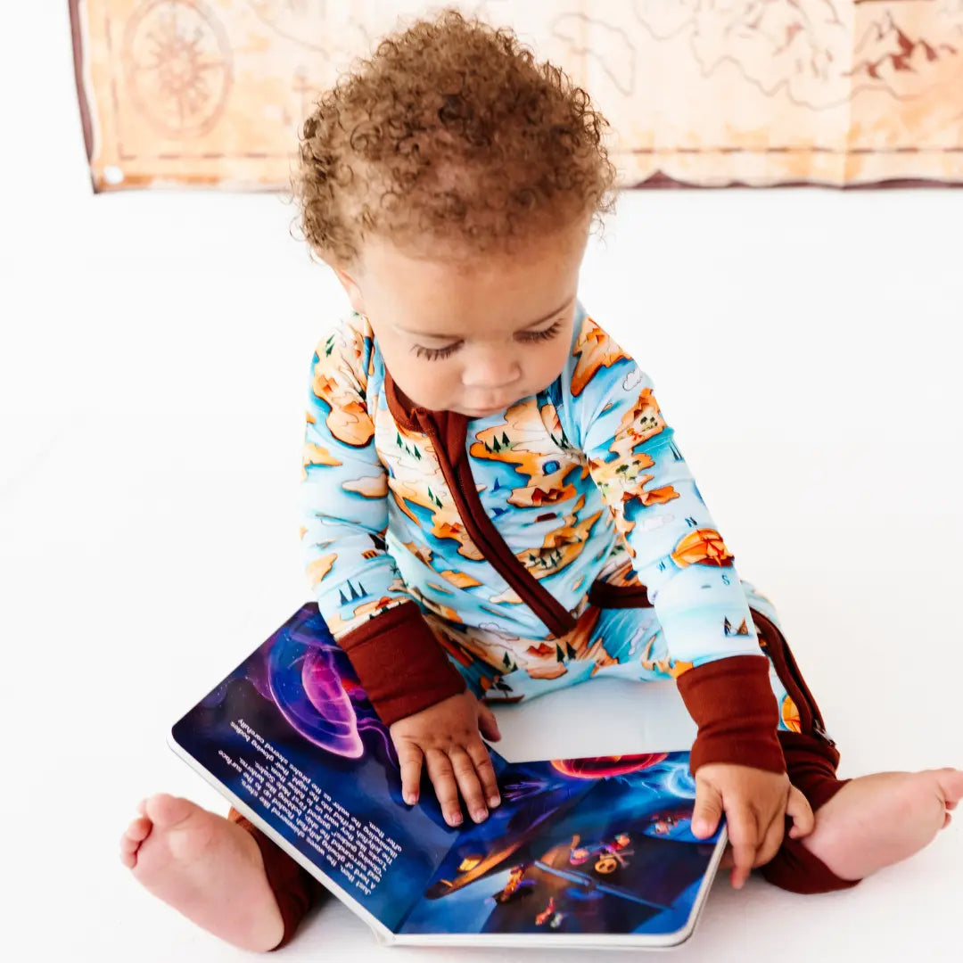 Curly-haired toddler sitting on the floor reading a colorful storybook, wearing a light blue onesie with an orange map and island print and brown trim, with a treasure-map decoration on the wall behind