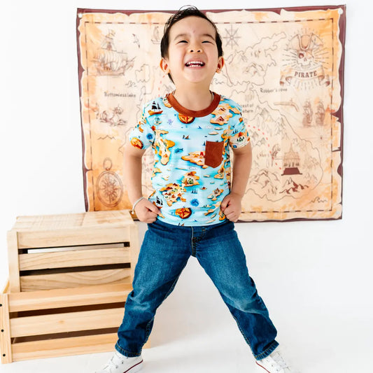 Child wearing a colorful shirt with a map design, standing in front of a pirate-themed wall hanging.