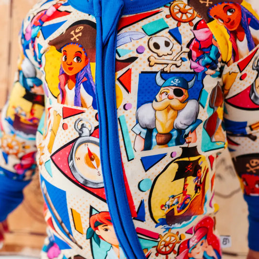 Close-up of a colorful pirate-themed onesie fabric with a blue zipper, showing cartoon pirate characters, a skull and crossbones, a compass, and a ship