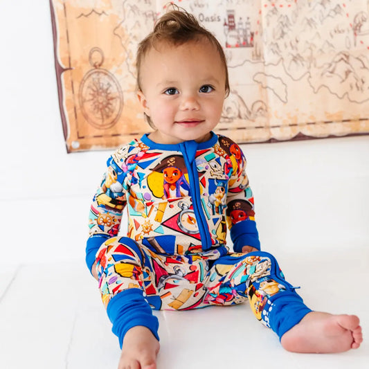 Smiling baby sitting on a white floor wearing a colorful pirate-themed cartoon-character onesie with blue cuffs, with a treasure map decoration on the wall behind