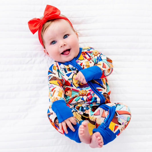 Smiling baby lying on a white blanket wearing a colorful pirate-themed zip-up onesie with blue cuffs and a red bow headband
