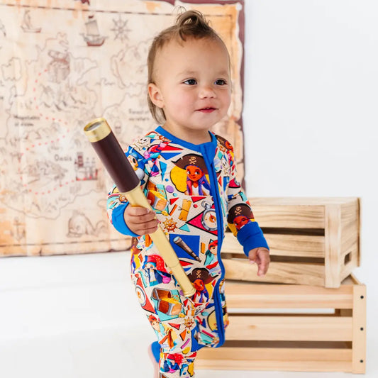 Toddler standing and holding a toy telescope, wearing a colorful pirate-themed onesie with blue cuffs, next to wooden crates, with a treasure map decoration on the wall behind