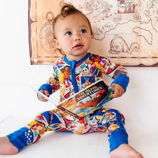 Toddler sitting on the floor holding a children's book titled The Island Adventures, wearing a colorful patterned onesie with blue cuffs, with a treasure map decoration on the wall behind