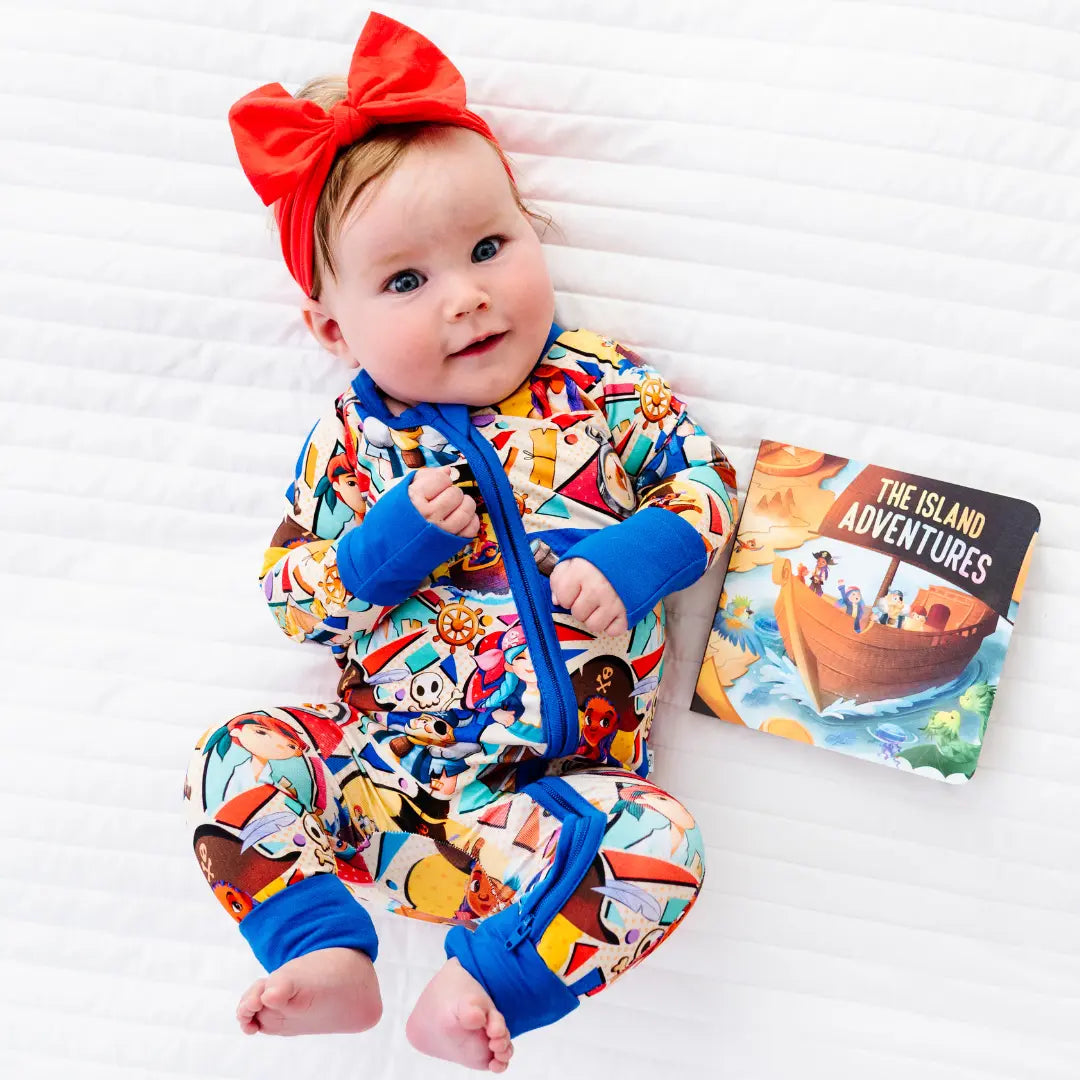 Baby lying on a white blanket wearing a colorful pirate-themed zip-up onesie with blue cuffs and a red bow headband, next to a children's book titled The Island Adventures