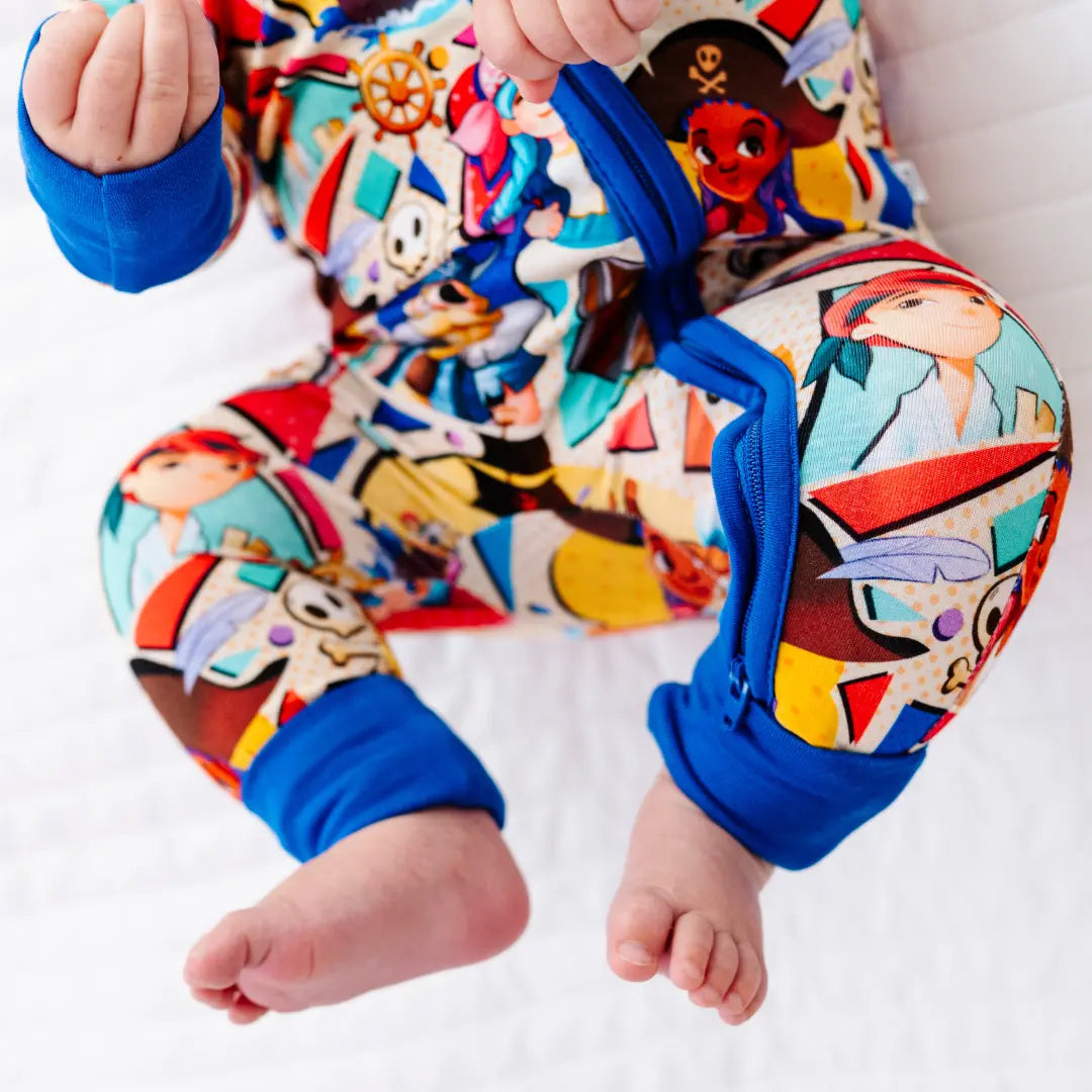 Close-up of a baby's legs and feet on a white blanket, wearing a colorful pirate-themed onesie with blue cuffs showing cartoon pirate characters and a skull