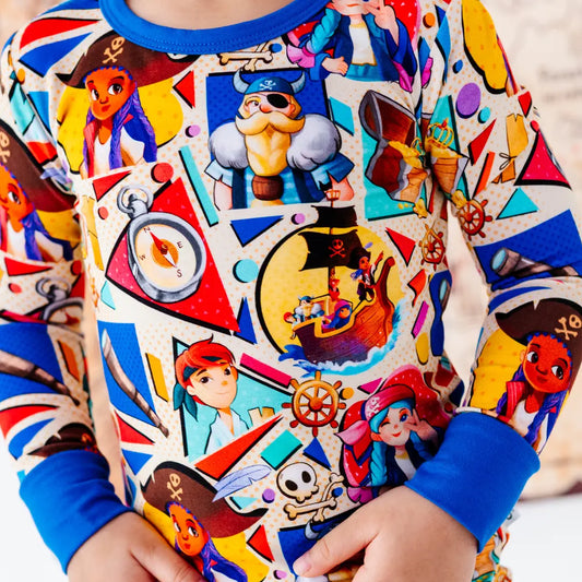 Close-up of a colorful pirate-themed pajama top with blue cuffs and collar, showing cartoon pirate characters, a compass, a ship's wheel, and a skull and crossbones, worn by a child