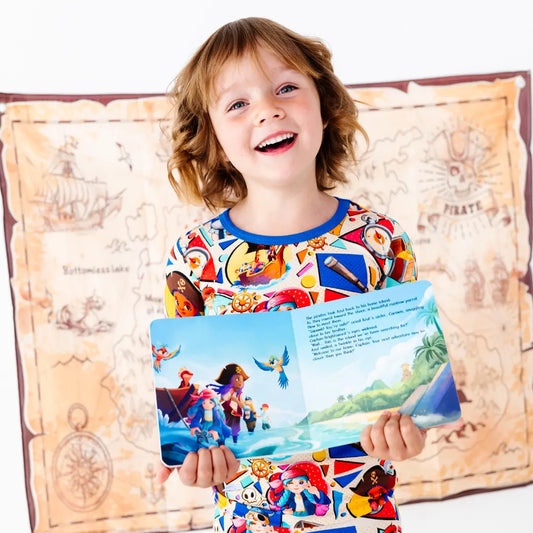 Laughing young girl holding open a children's picture book showing a pirate and island scene, wearing a colorful pirate-themed pajama top with a blue collar, with a treasure map decoration on the wall behind