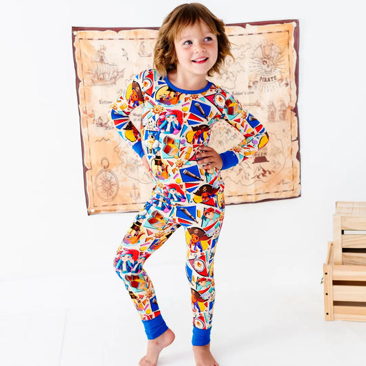Smiling young girl standing with a hand on her hip, wearing a colorful pirate-themed pajama set with blue cuffs and collar, next to wooden crates, with a treasure map decoration on the wall behind