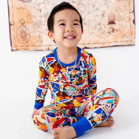 Smiling young boy sitting cross-legged on a white floor, wearing a colorful pirate-themed cartoon-character pajama set with blue cuffs, with a treasure map decoration on the wall behind