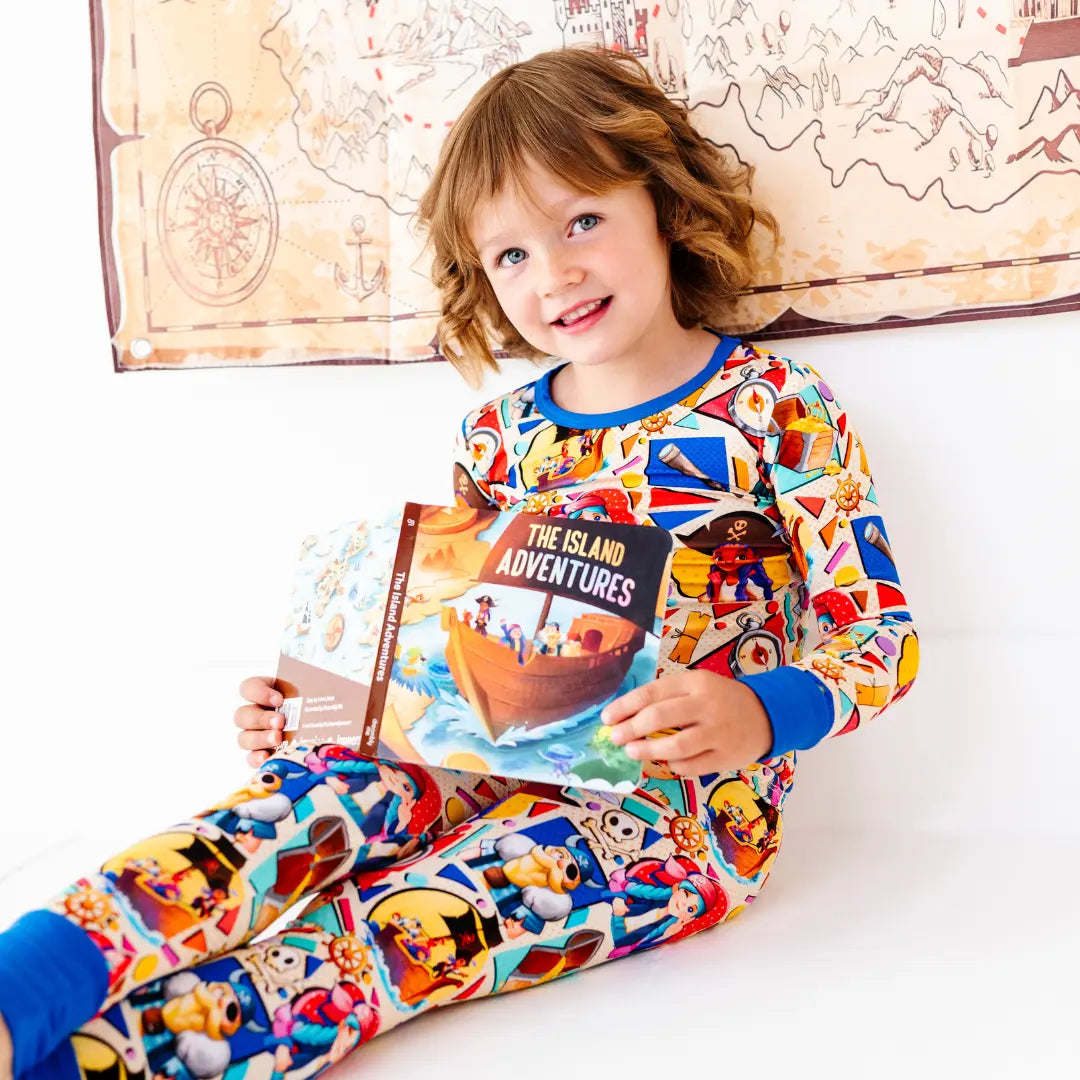 Smiling young girl sitting and holding a children's book titled The Island Adventures, wearing a colorful pirate-themed pajama set with blue cuffs, with a treasure map decoration on the wall behind