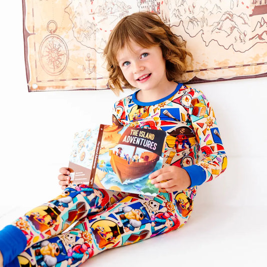 Smiling young girl sitting and holding a children's book titled The Island Adventures, wearing a colorful pirate-themed pajama set with blue cuffs, with a treasure map decoration on the wall behind