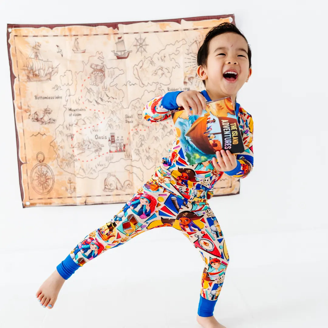 Laughing young boy in a playful stance holding a children's book titled The Island Adventures, wearing a colorful pirate-themed cartoon-character pajama set with blue cuffs, with a treasure map decoration on the wall behind