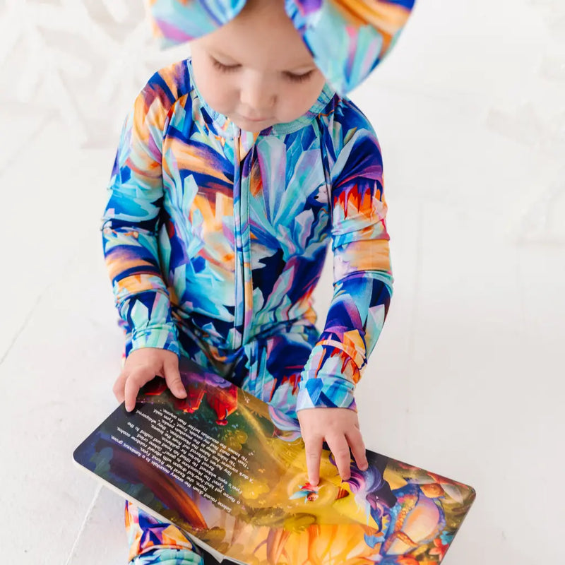 Child wearing a colorful onesie and hat, holding a book with a matching design.