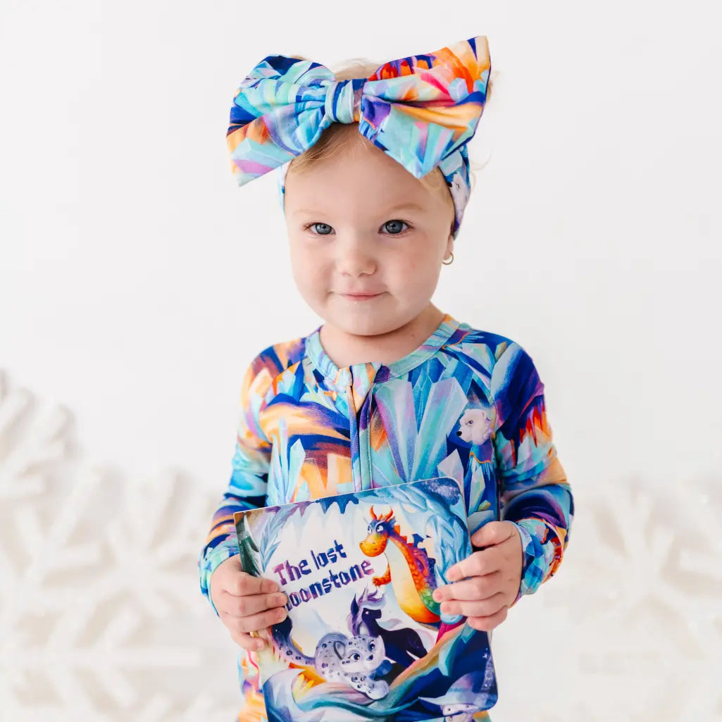 Child wearing a colorful outfit and headband, holding a book with a rainbow design.