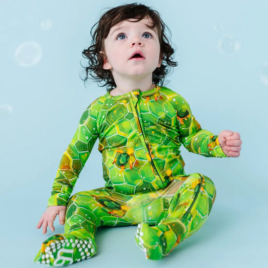A baby wearing a green one-piece zipper pajama with geometric sea turtle patterns, sitting on a blue background.