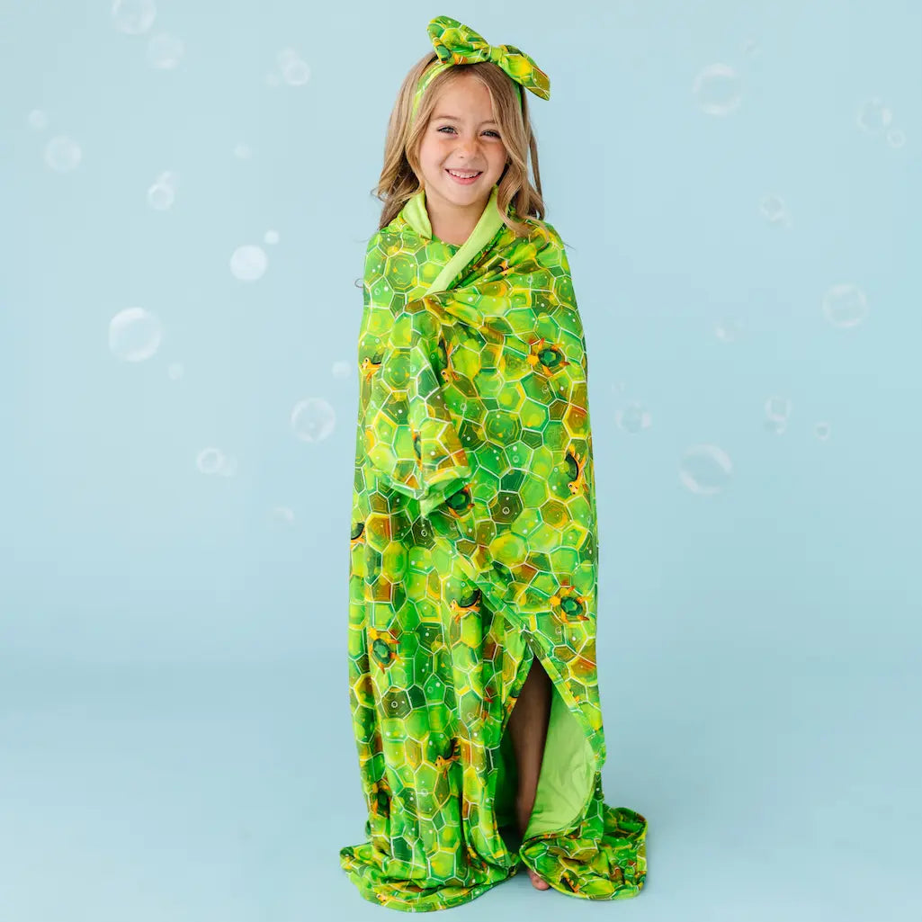 A child wrapped in a green blanket with a geometric turtle pattern, standing against a blue background with bubbles around.