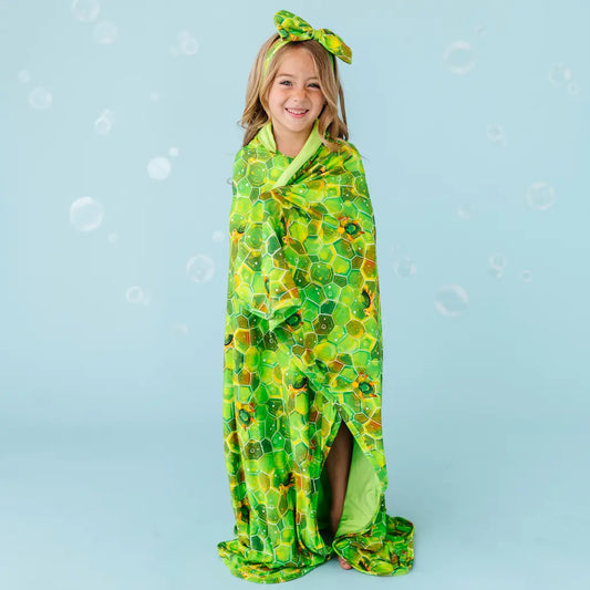 A child wrapped in a green blanket with a geometric turtle pattern, standing against a blue background with bubbles around.