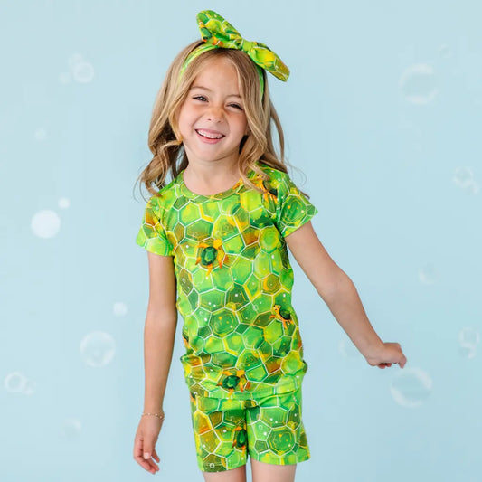 A child wearing a short pajama set with a geometric green shell patterns and hexagonal designs, complemented by a headband with a similar design.