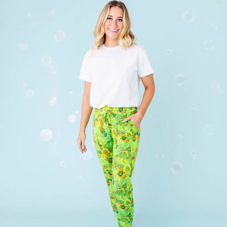 A woman standing and smiling, wearing white shirt and green pajama pants with a geometric turtle pattern.