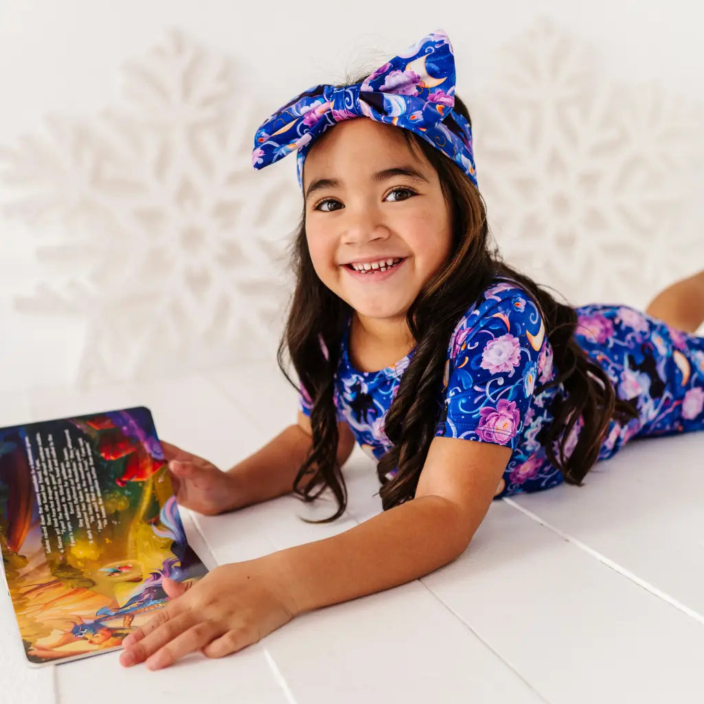 Young girl in a floral outfit reading a book on a white floor.