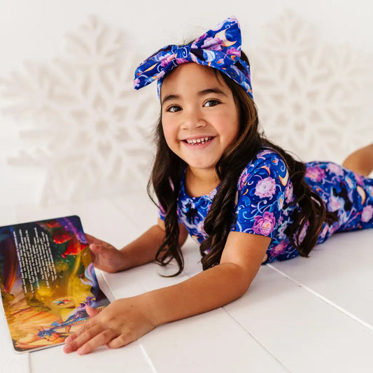 Young girl in a floral outfit reading a book on a white floor.