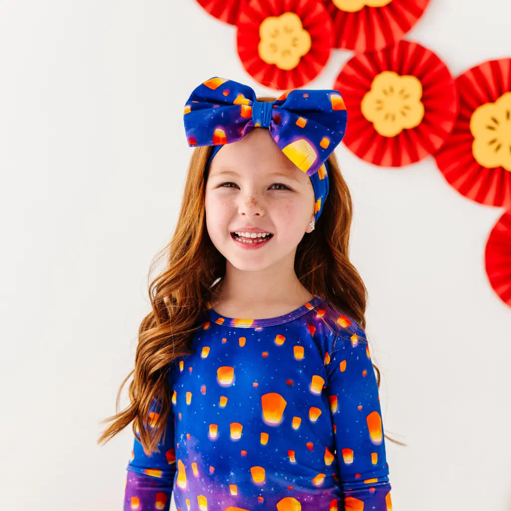 Young girl wearing a blue shirt with candy corn pattern and colorful headband, standing in front of red and yellow decorative fans.