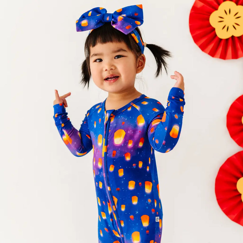 Child wearing a colorful outfit with a blue bow, standing against a white background with red decorative elements.