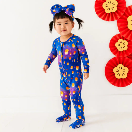 Child wearing a colorful onesie with abstract patterns, standing against a white background with decorative red and yellow fans.