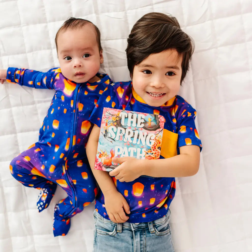 Two children wearing colorful outfits lying on a white blanket, with one holding a book.