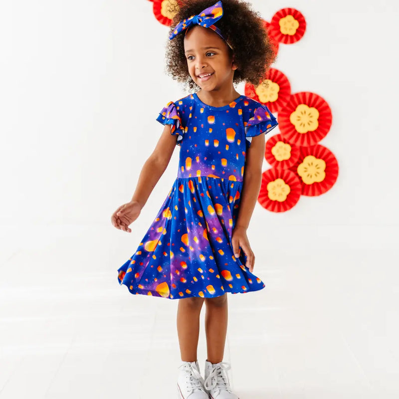 Child wearing a blue dress with orange polka dots, standing in front of decorative flowers.