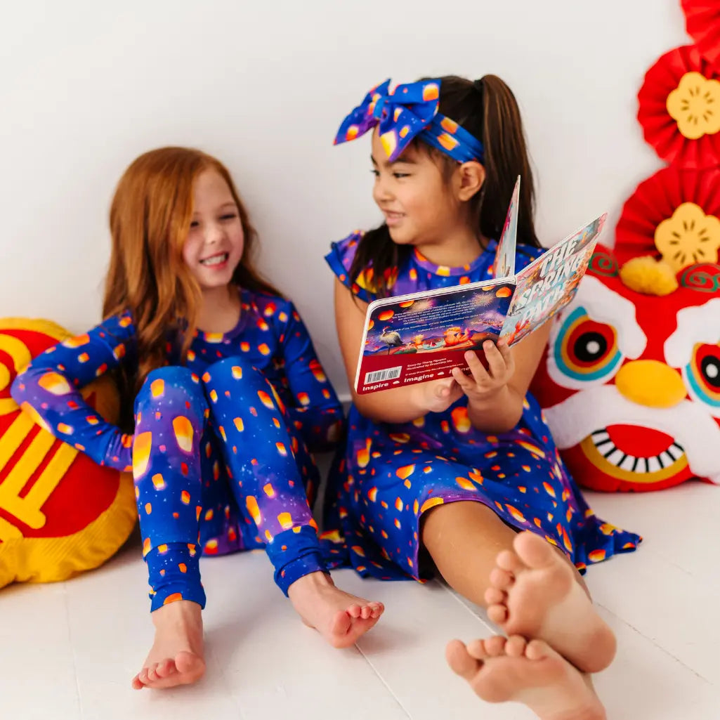 Two children in colorful outfits sitting on a white floor with a red and yellow decorative item in the background.