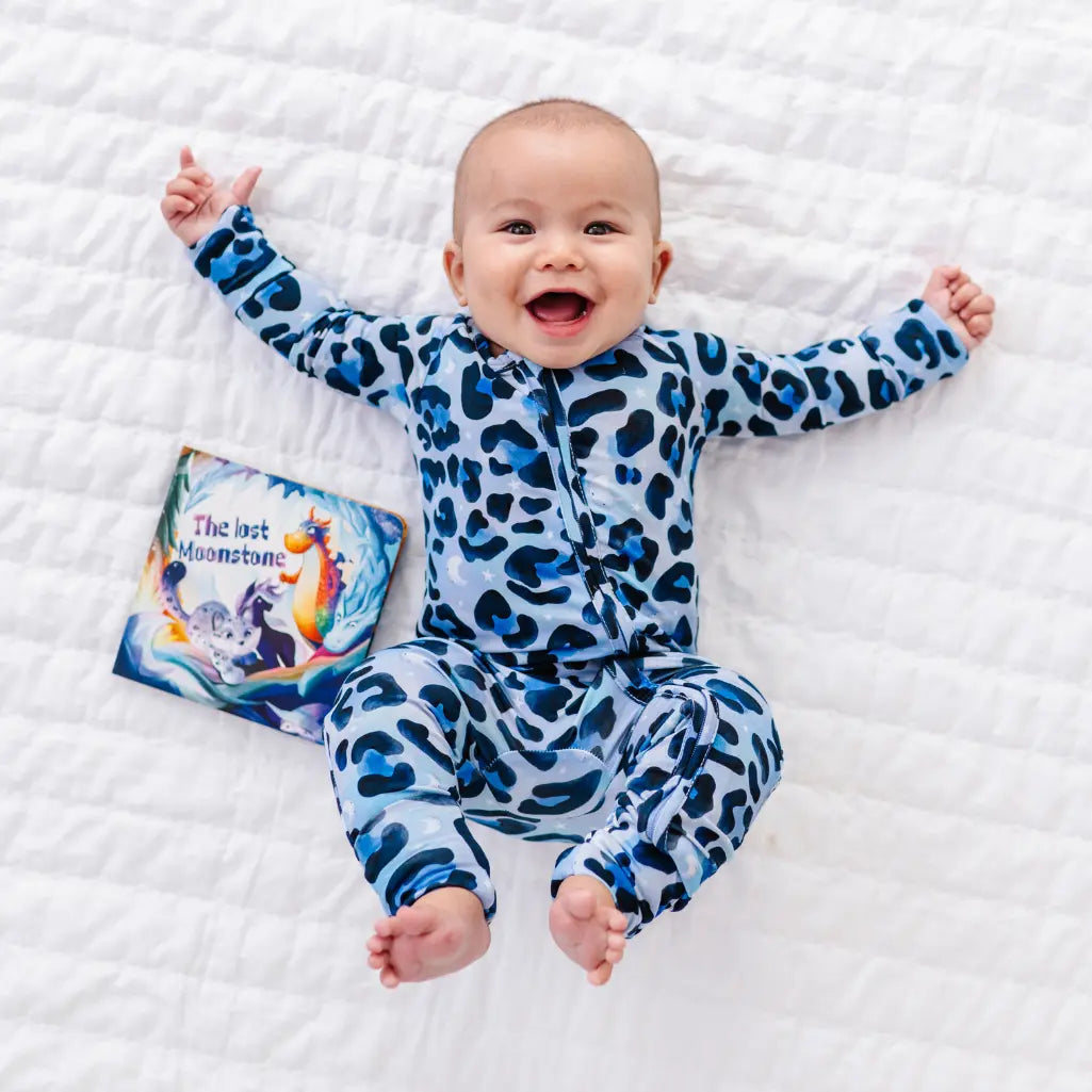 Baby wearing a blue leopard print onesie on a white blanket with a book titled 'The Last Moonstone'.