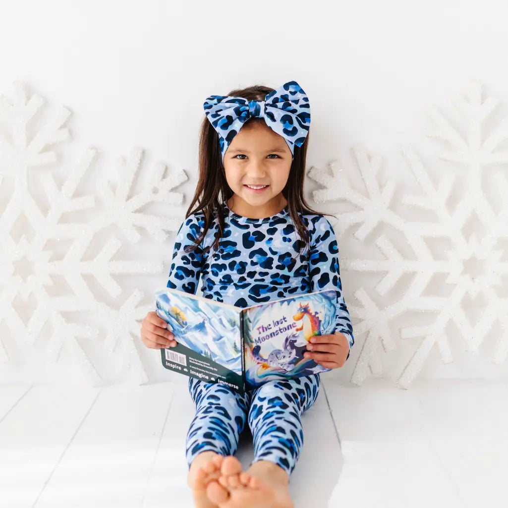 Child wearing a blue leopard print outfit with matching headband, sitting on a white surface holding books.