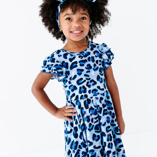 Smiling young girl standing with a hand on her hip, wearing a blue leopard-print ruffle-sleeve dress and a matching bow headband, on a white background