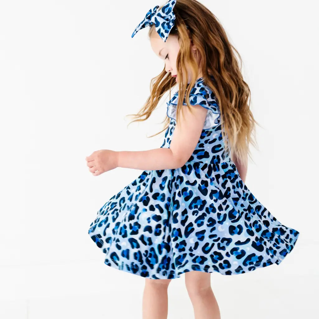 Young girl twirling in a blue leopard-print ruffle dress with a matching bow headband, on a white background