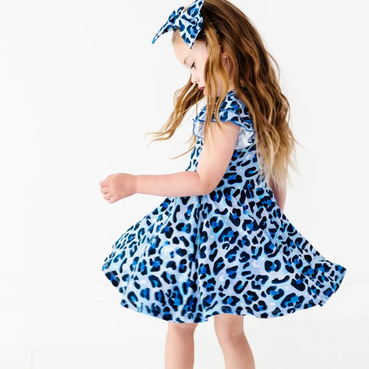 Young girl twirling in a blue leopard-print ruffle dress with a matching bow headband, on a white background