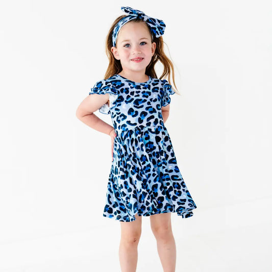 Smiling young girl standing with a hand on her hip, wearing a blue leopard-print ruffle-sleeve dress and a matching bow headband, on a white background